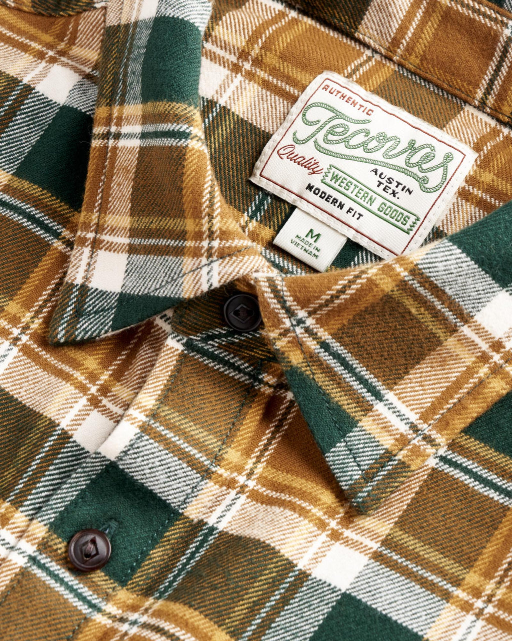 Close-up of a green, brown, and white plaid flannel shirt with dark buttons and a sewn label reading “Jeans Authentic Quality Western Goods” and “Modern Fit.”.