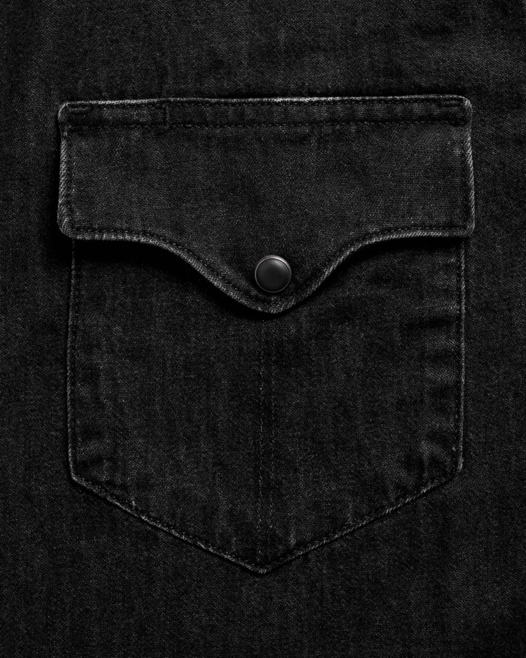Close-up of a black denim shirt pocket with a flap and a black button in the center. The fabric has a subtle texture and stitching is visible around the edges.