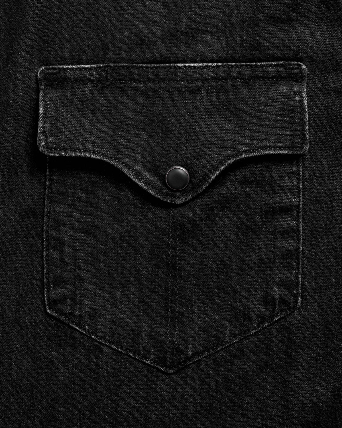 Close-up of a black denim shirt pocket with a flap and a black button in the center. The fabric has a subtle texture and stitching is visible around the edges.