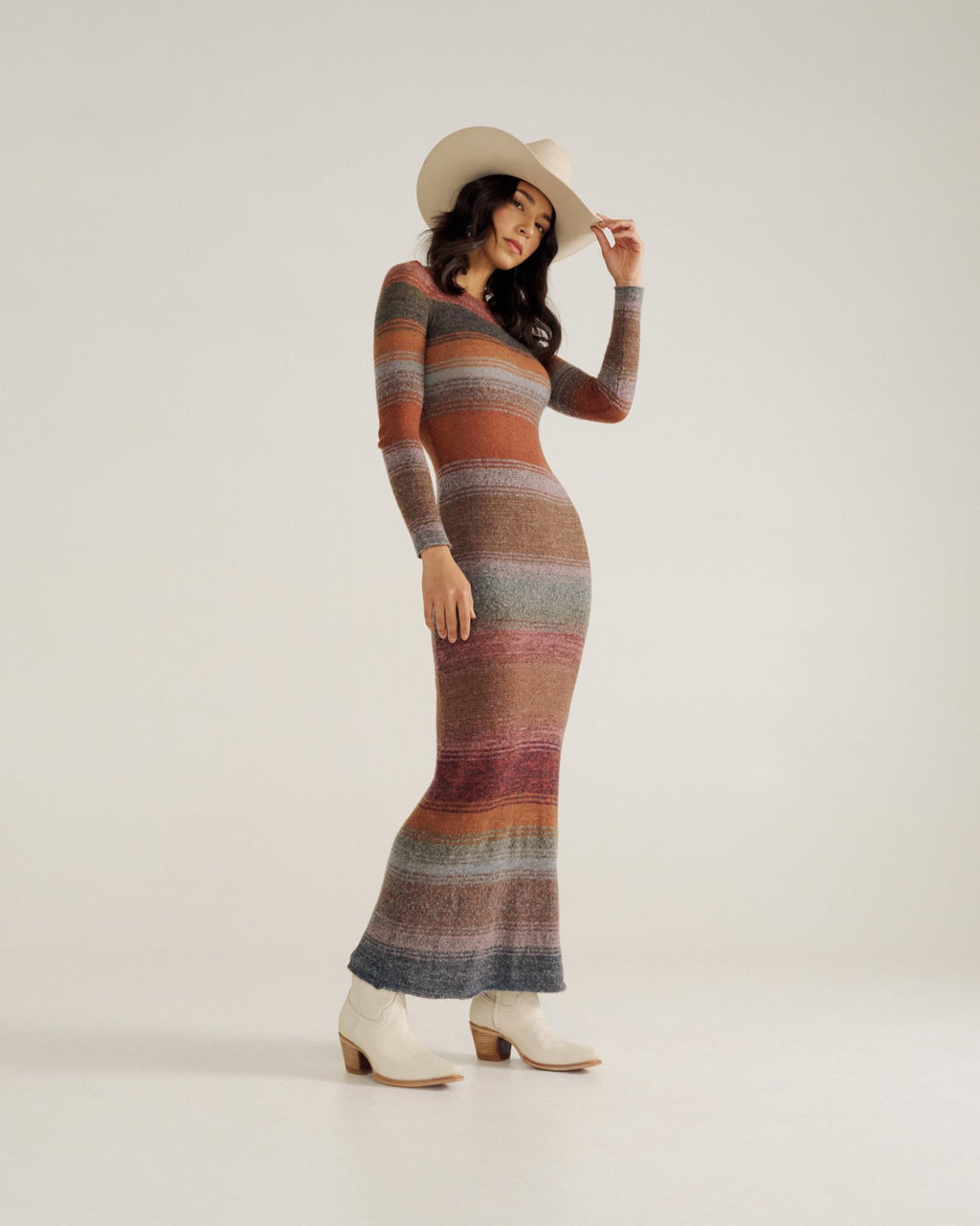 Woman wearing a striped, long-sleeve, fitted maxi dress, cream cowboy hat, and cream ankle boots standing against a plain background.