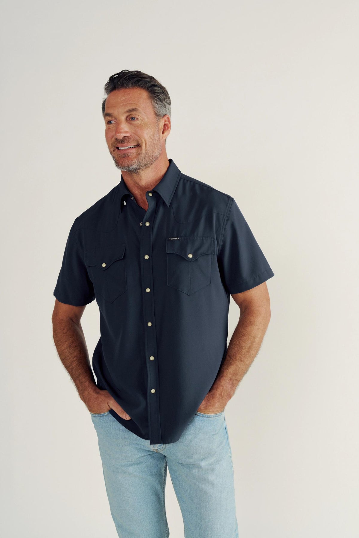 A man with short hair and a beard wears a navy short-sleeve button-up shirt and light blue jeans, standing against a plain off-white background.
