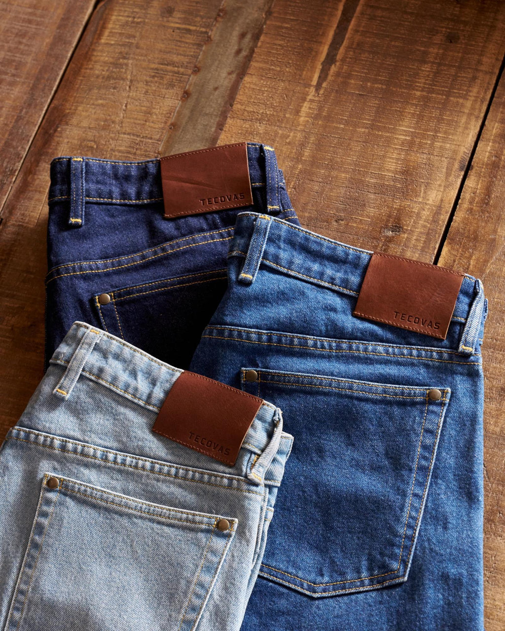 Three pairs of denim jeans in light, medium, and dark blue washes with brown leather brand patches, laid out on a wooden surface.