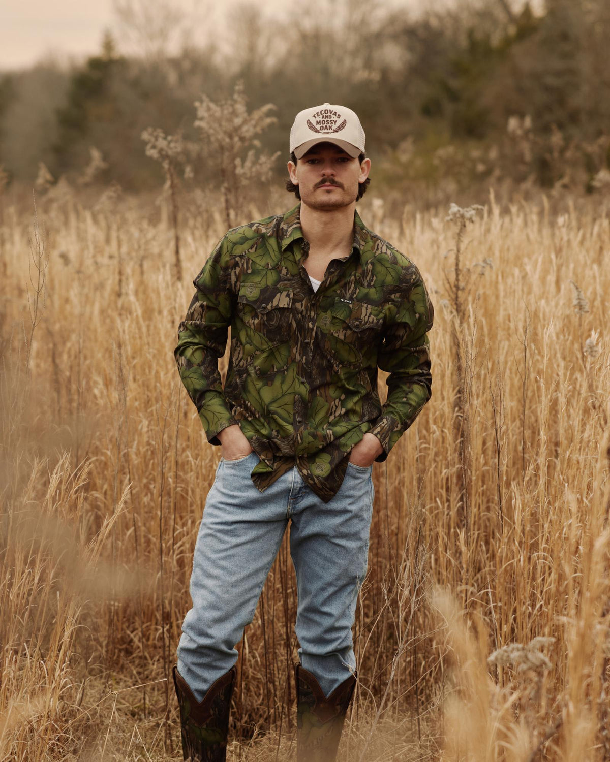 Man standing in a field wearing a camo shirt, light-wash jeans, dark cowboy boots, and a Tecovas x Mossy Oak trucker hat.