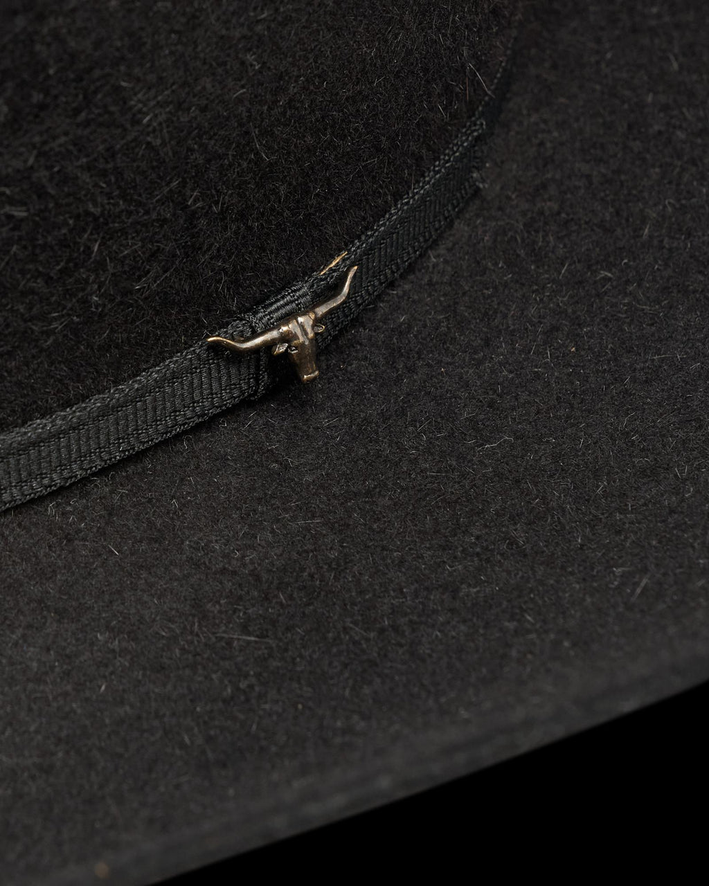 Close-up of a black felt hat with a black ribbon band and a small metallic longhorn ornament attached to the band.