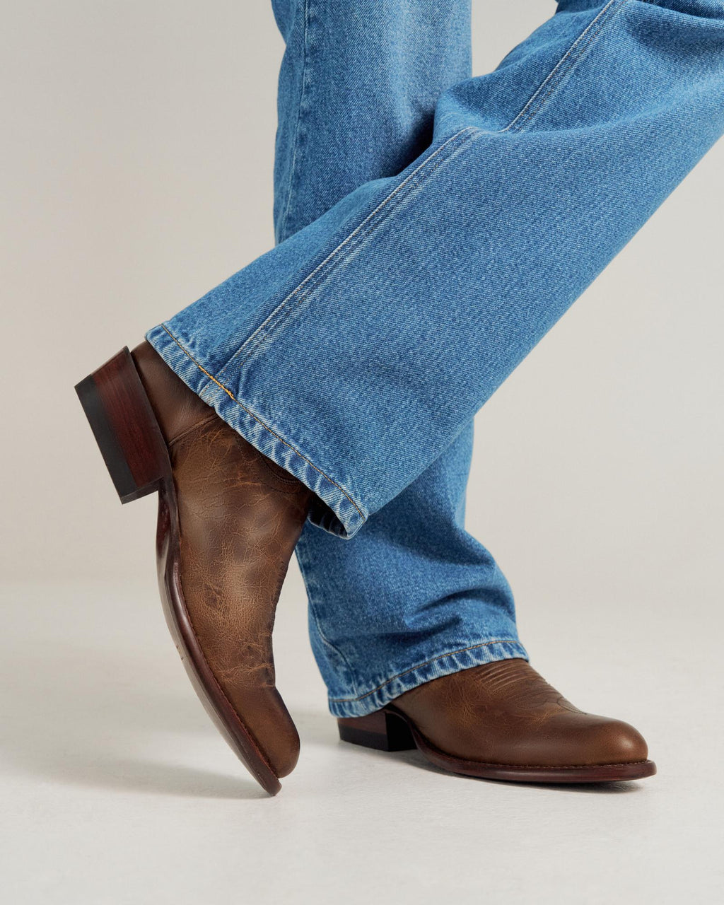 A person wearing light blue jeans and brown leather boots stands with one leg crossed over the other against a plain background.