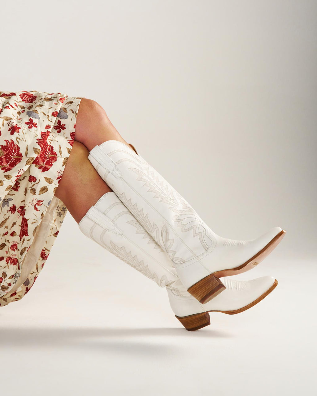 The Abby Wide Calf in Snow Cowhide. Person wearing a floral skirt and Snow cowhide white cowboy boots with brown heels; legs crossed against a plain light background.