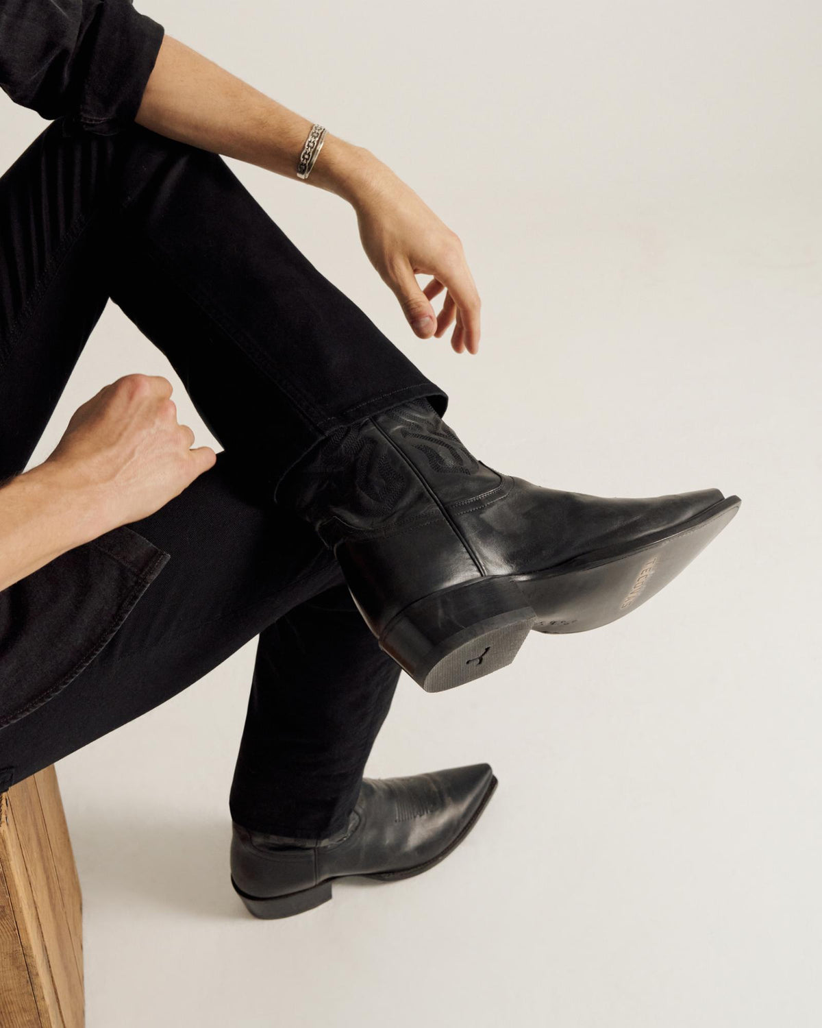 Person seated on a wooden block wears black jeans, a silver bracelet, and black pointed-toe boots, with one leg crossed over the other against a plain background.