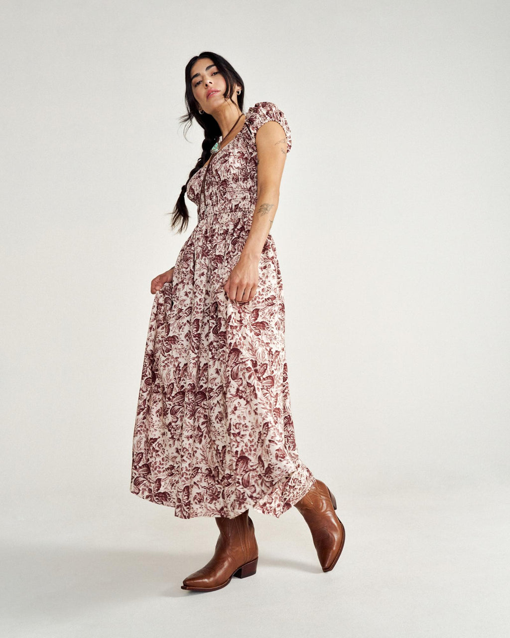 A person wearing a patterned long dress and brown cowboy boots stands against a plain background.