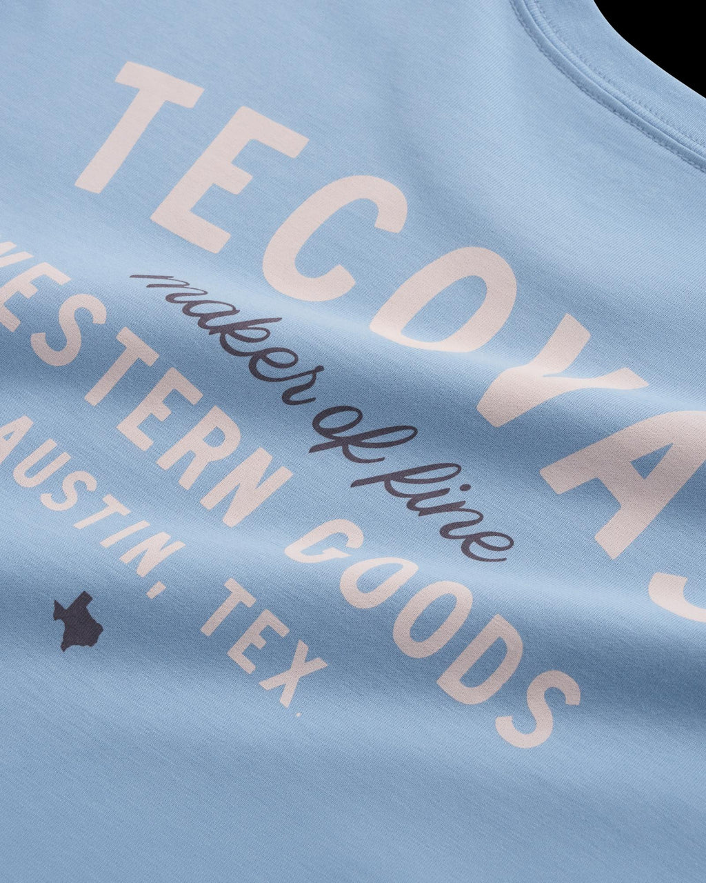 Close-up of a light blue shirt with white text reading "TECDYAS maker of fine WESTERN GOODS AUSTIN, TEX," plus a small Texas state outline icon.