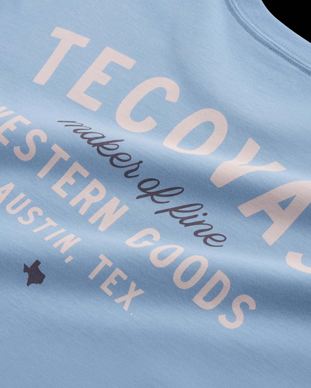 Close-up of a light blue shirt with white text reading "TECDYAS maker of fine WESTERN GOODS AUSTIN, TEX," plus a small Texas state outline icon.
