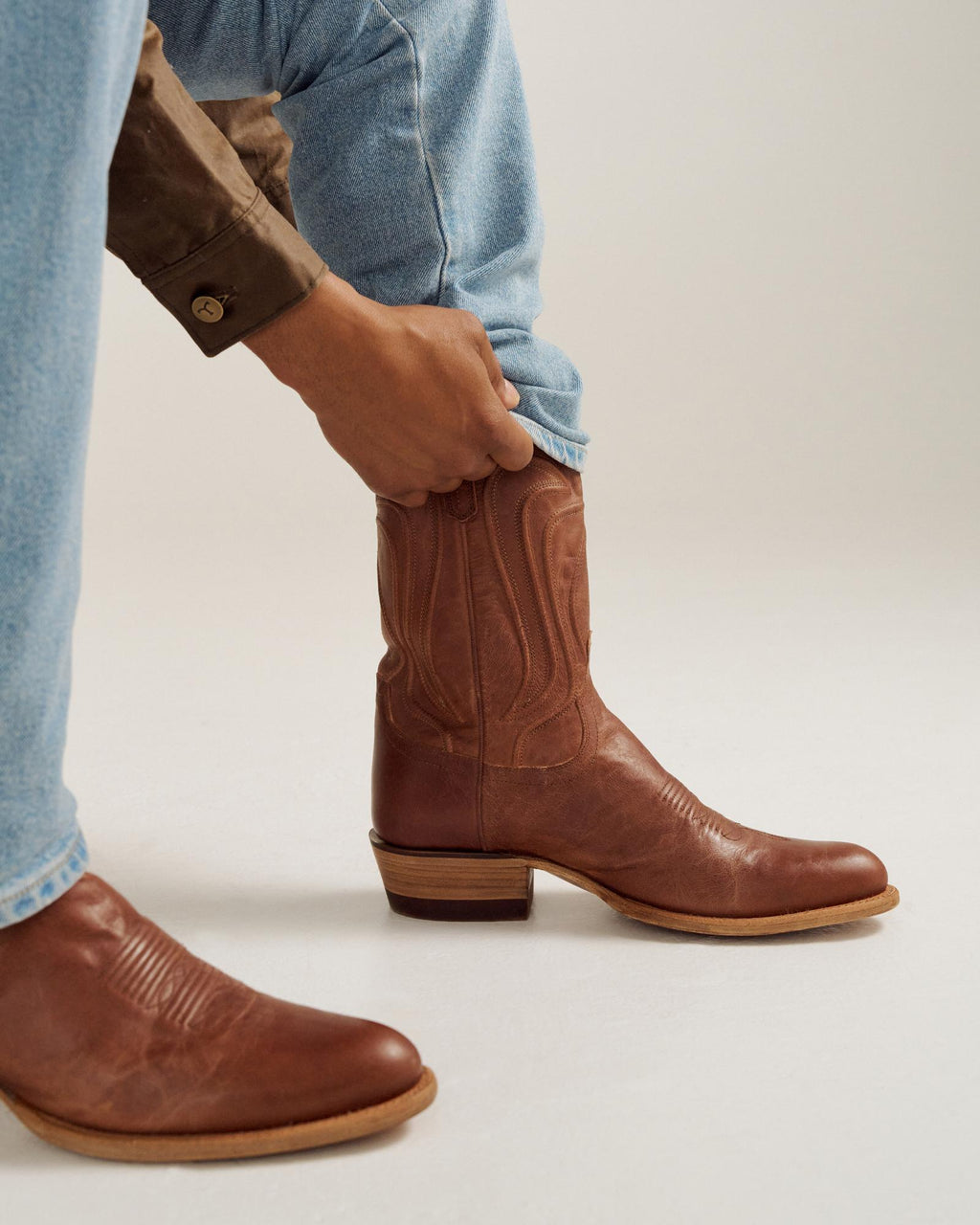 A person wearing light blue jeans and a brown shirt adjusts a brown leather cowboy boot on their left foot.
