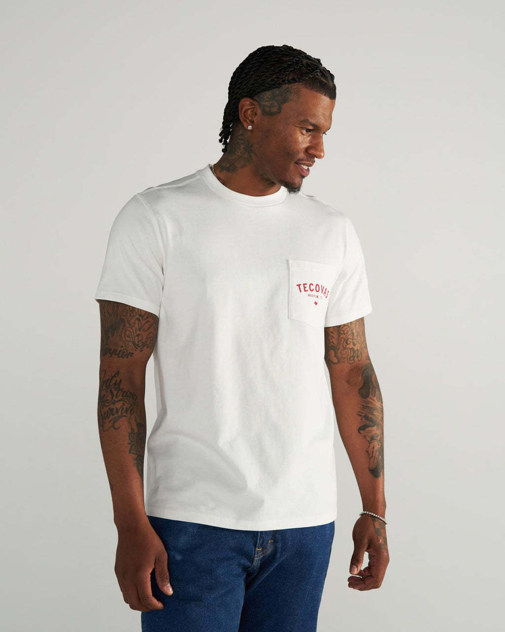 Man wearing a white T-shirt with “TECOVAS” in red on the pocket, blue jeans, and displaying tattoos on both arms, standing against a plain light background.