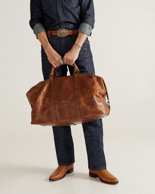 Person wearing denim outfit and cowboy boots holding a large brown leather duffel bag by its handles, standing against a plain background.