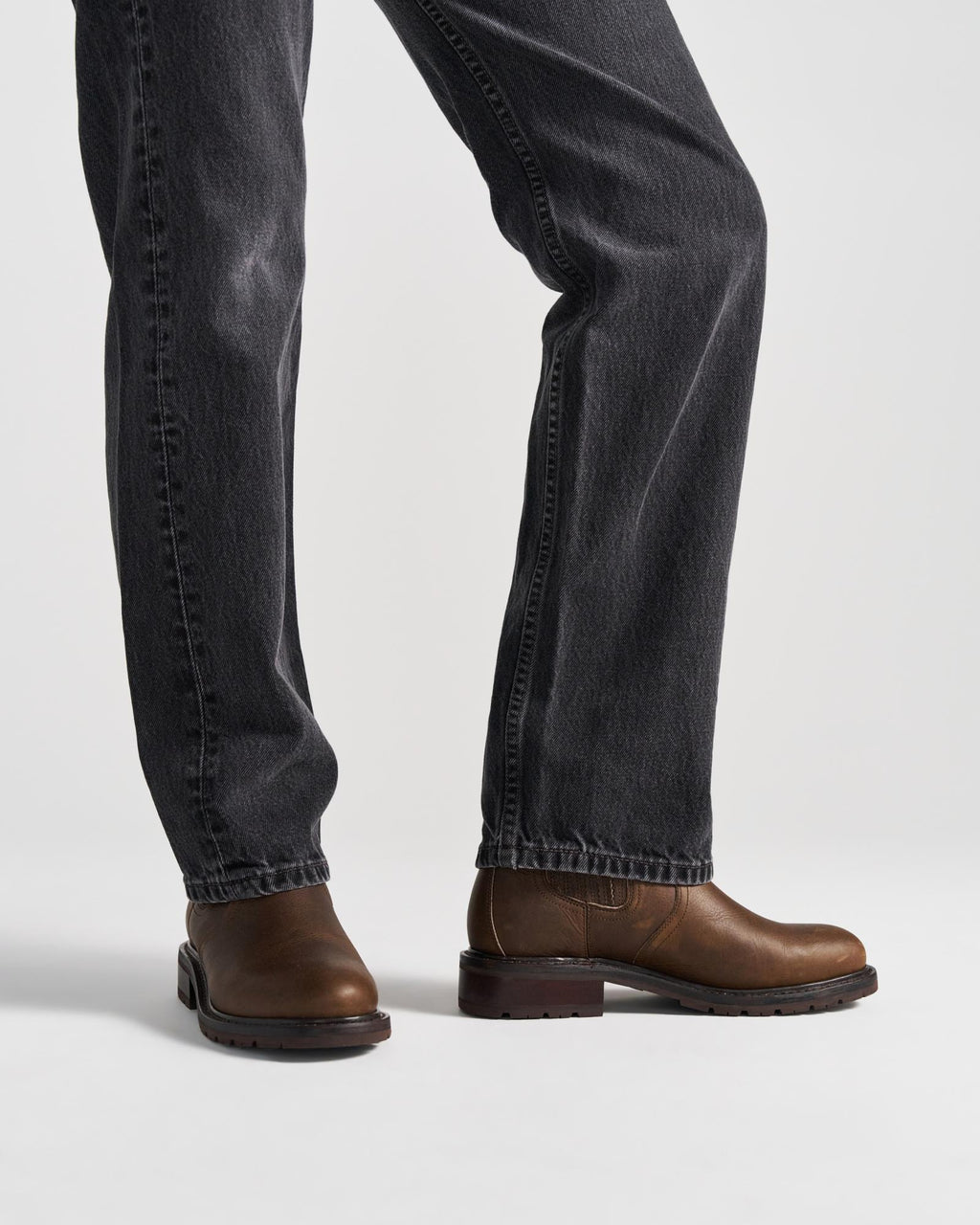 The Chelsea in Cafe Cowhide. Person wearing dark jeans and brown leather Cafe Cowhide shoes, standing on a white background. Only legs and feet are visible.