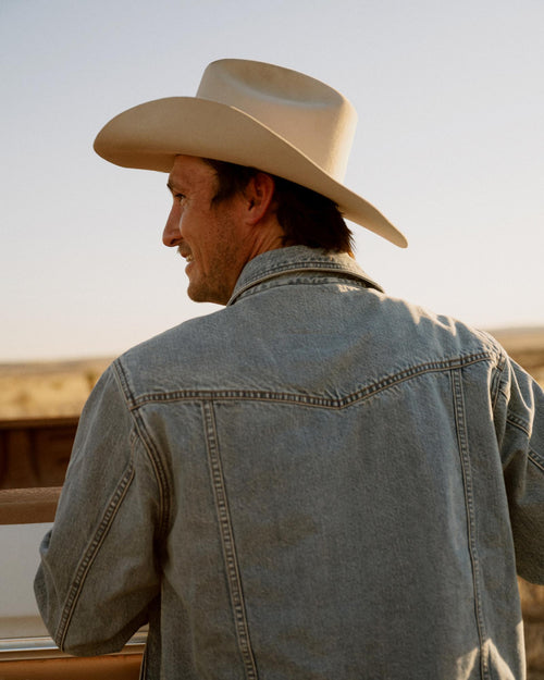A man wearing a cowboy hat and denim jacket stands outdoors, facing away and looking toward an open, sunlit landscape.
