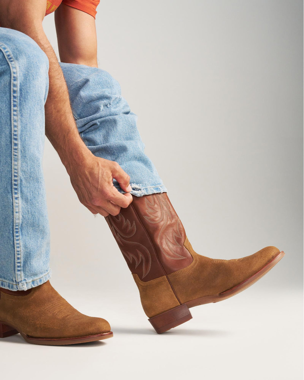 The Buck in Copper Cowhide. A person in blue jeans and an orange shirt tucks their pant leg into a sturdy Copper Cowhide cowboy boot.