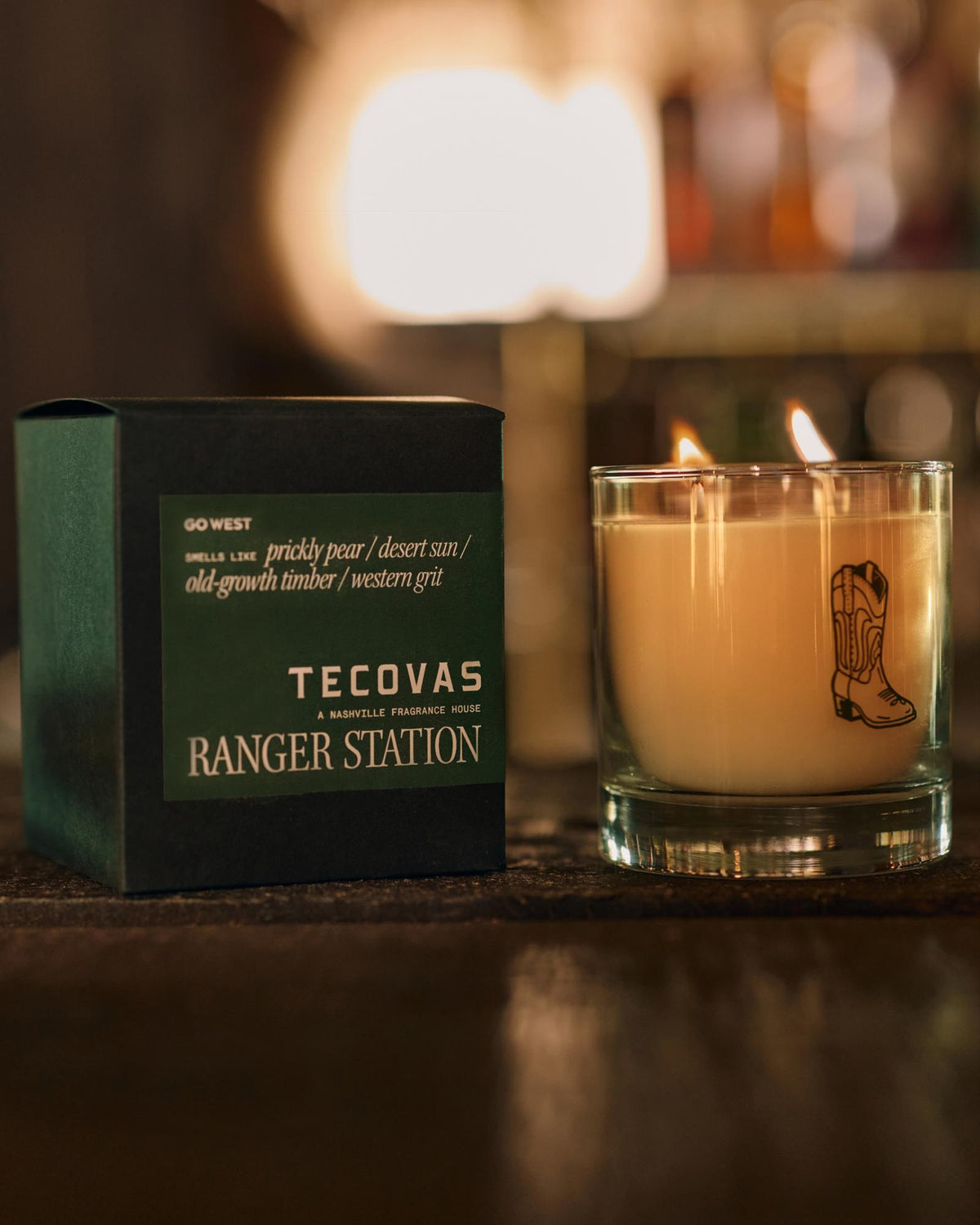 A lit candle with a boot design beside a dark green box labeled "Tecovas Ranger Station" on a wooden surface.