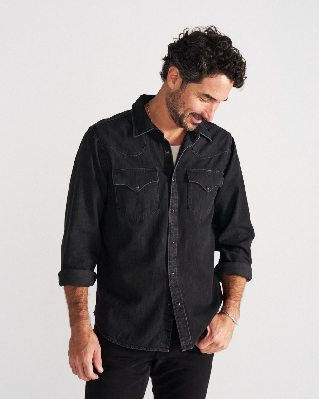 Man with curly hair and a beard, wearing a black denim button-up shirt with rolled sleeves, stands against a plain white background, looking down and slightly smiling.