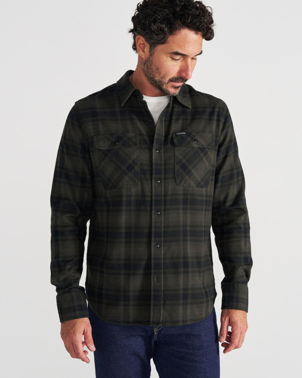 A man stands against a plain background, wearing a dark green and black plaid button-up shirt over a white t-shirt and blue jeans, looking down and to his left.