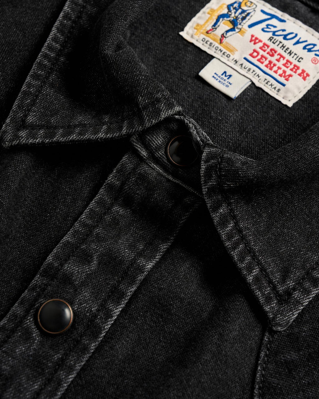 Close-up of a black denim shirt with metal snap buttons and a visible Tecovas Western Denim label on the collar.