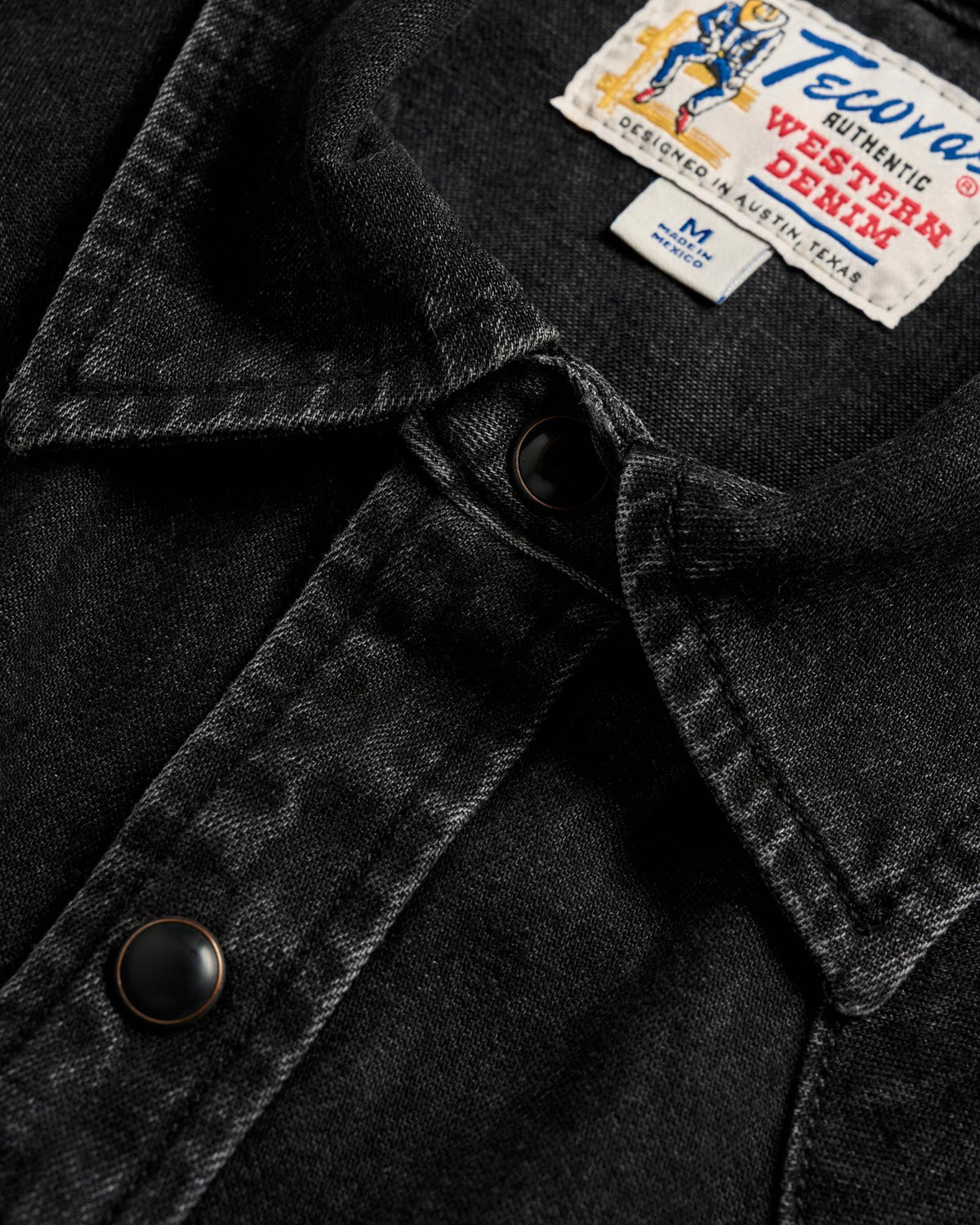 Close-up of a black denim shirt with metal snap buttons and a visible Tecovas Western Denim label on the collar.