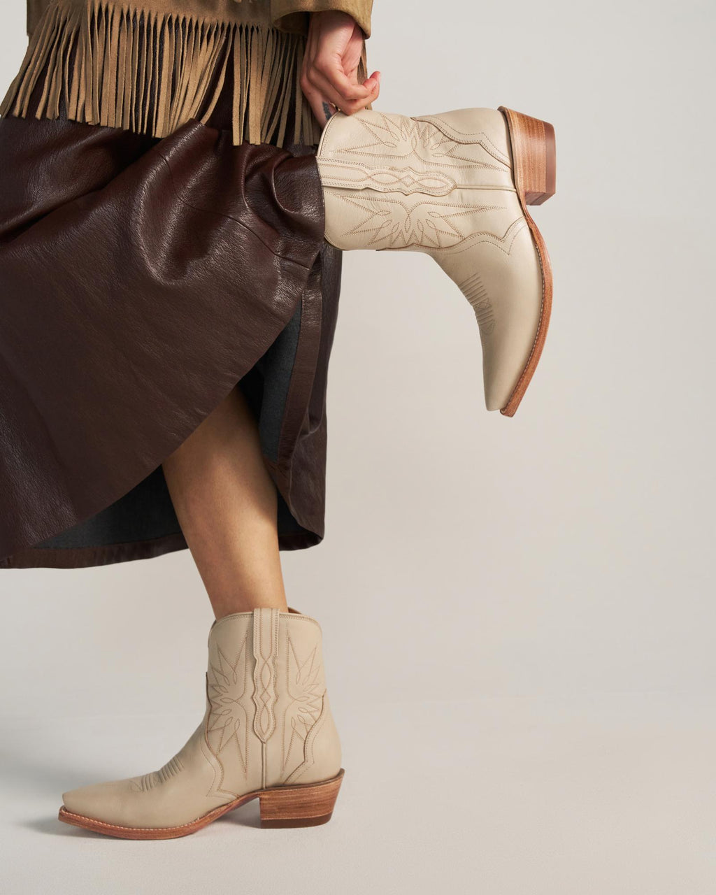 A person wearing a dark brown faux leather skirt, a tan fringe jacket, and light tan embroidered cowboy boots with wooden heels inspired by The Lynn Cowhide in a soft cream shade.
