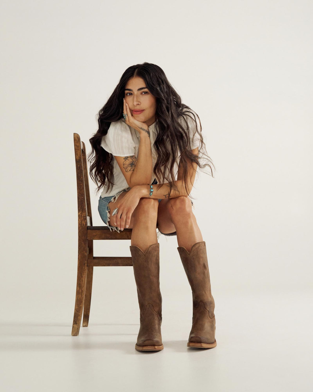 A woman with long dark hair sits on a wooden chair at The Charlie Goat cafe, resting her chin on her hand. She wears a white top, denim shorts, and brown cowboy boots against a plain background.