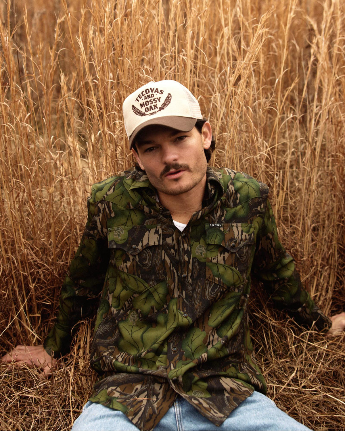 A person in a camouflage shirt and a tan cap sits in tall dry grass, looking toward the camera with a neutral expression.