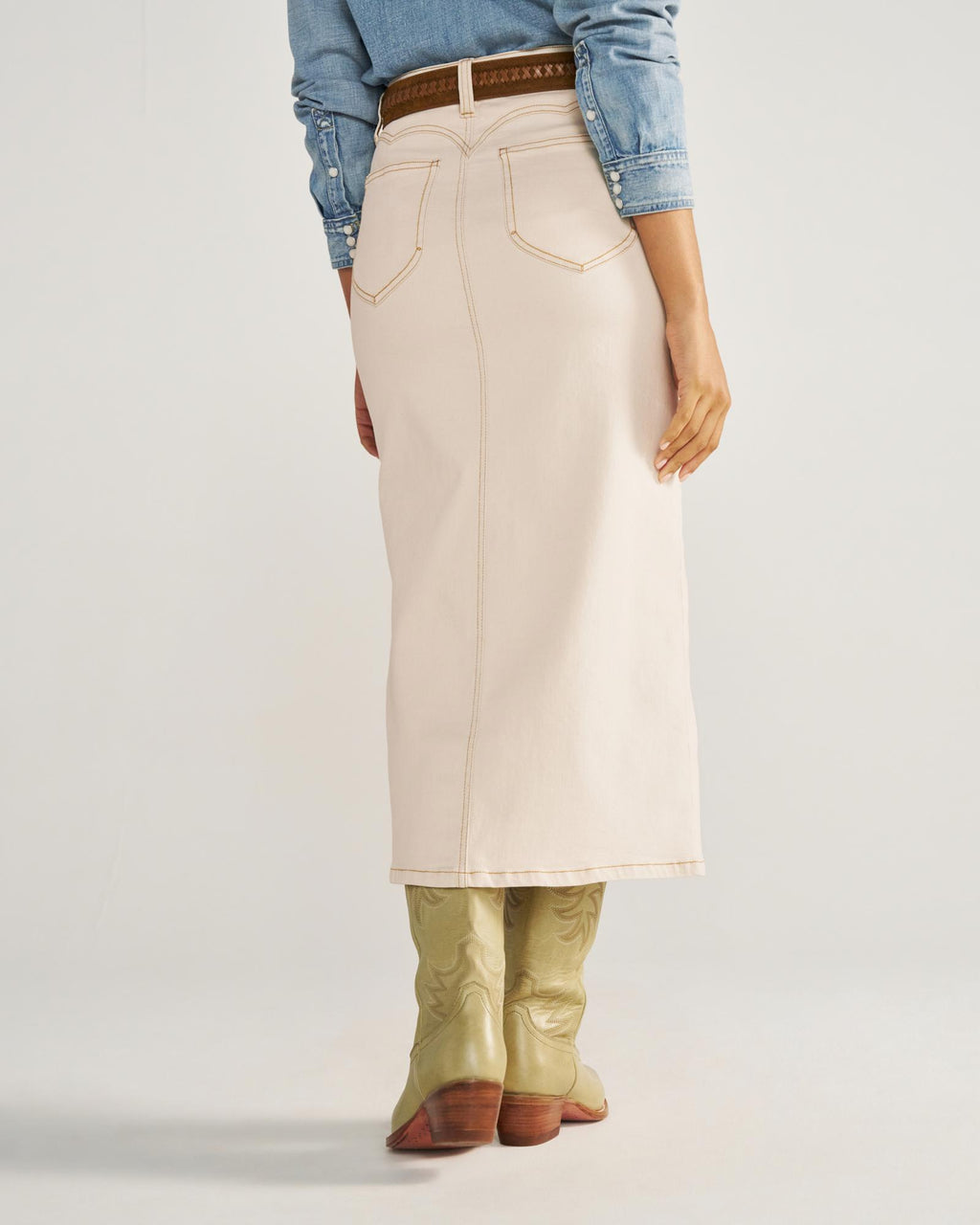 A person wearing a light denim skirt, a brown belt, a blue long-sleeve shirt, and light green cowboy boots, shown from the back.