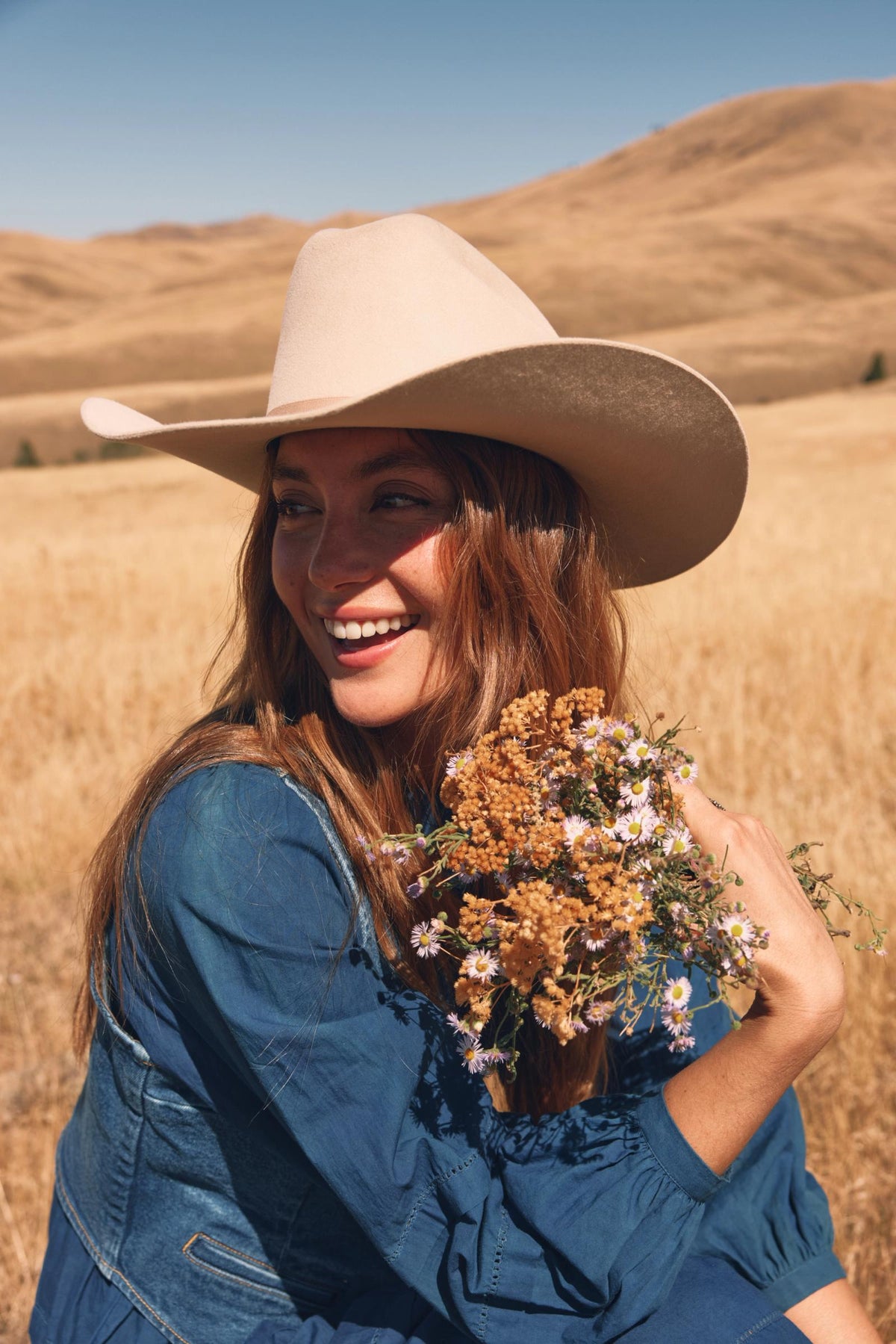 A person wearing a cowboy hat smiles while holding a bouquet of wildflowers in a dry, grassy field under a clear blue sky.