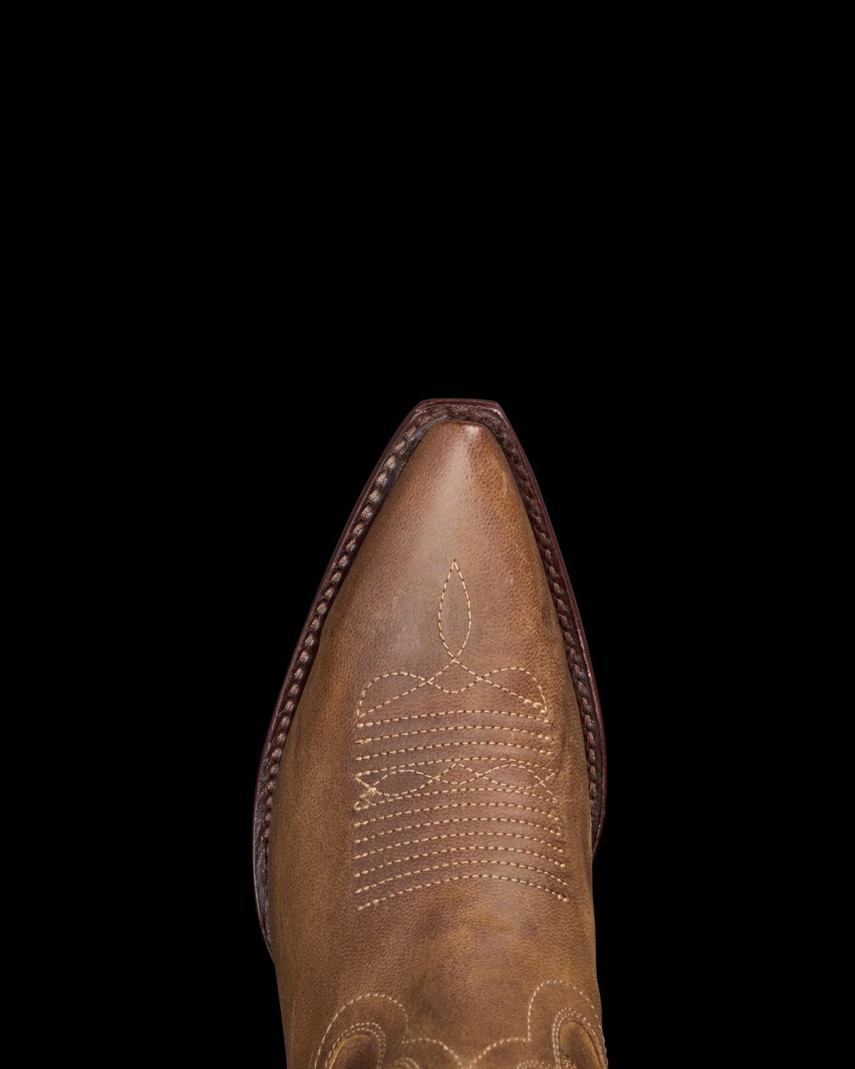 A close-up of a brown leather cowboy boot's pointed toe with intricate stitching on a black background.