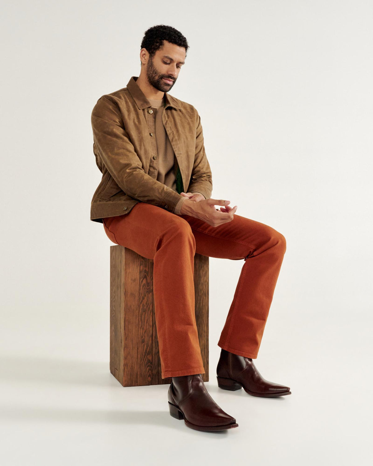 Man seated on a wooden block wearing a brown jacket, beige shirt, orange pants, and dark boots, looking at a small object in his hands against a plain background.