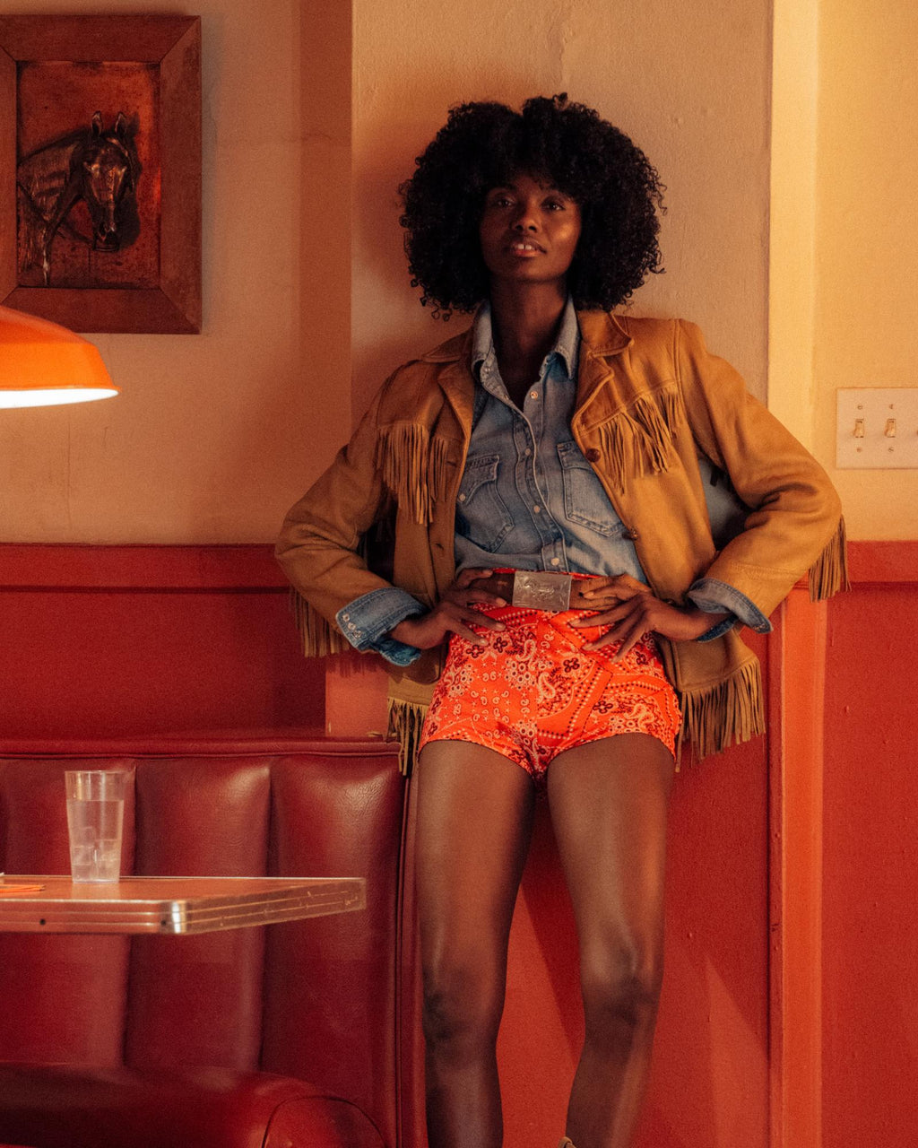 A person with curly hair stands in a retro diner, wearing a fringed jacket, denim shirt, and red patterned shorts, with hands on hips.