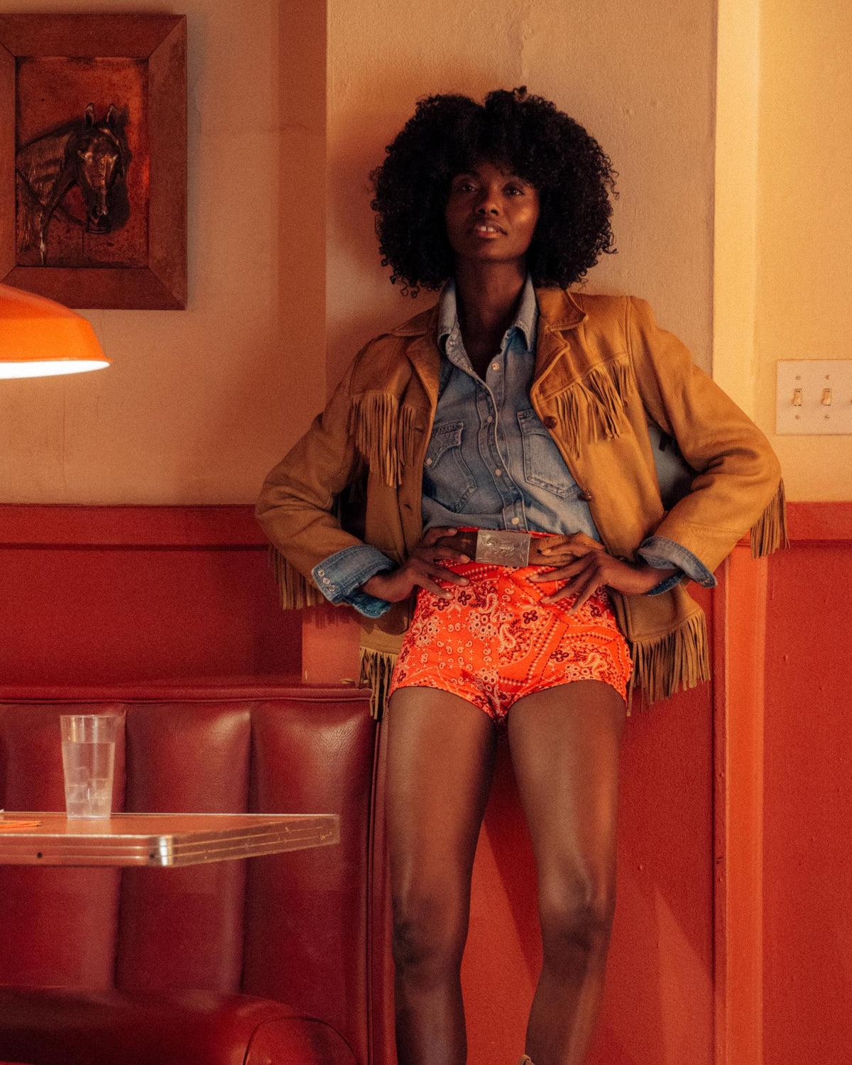 A person with curly hair stands in a retro diner, wearing a fringed jacket, denim shirt, and red patterned shorts, with hands on hips.