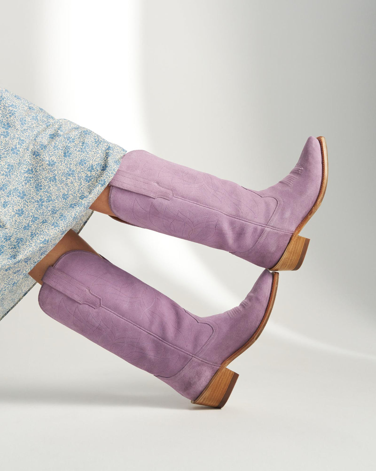A person wearing a light blue floral dress and The Annie Suede knee-high lavender boots with wooden heels, legs extended and crossed at the ankles.
