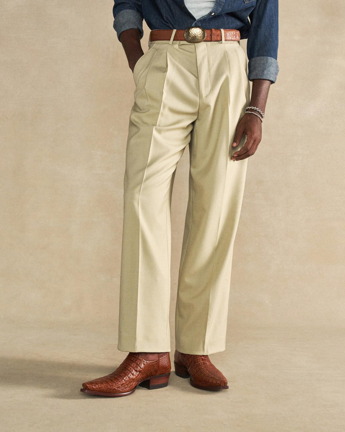 The Garrett in Pecan Hornback Caiman. A person wearing beige pleated dress pants, a brown pecan belt with a large buckle, brown Hornback Caiman cowboy boots, and a denim shirt with sleeves rolled up.