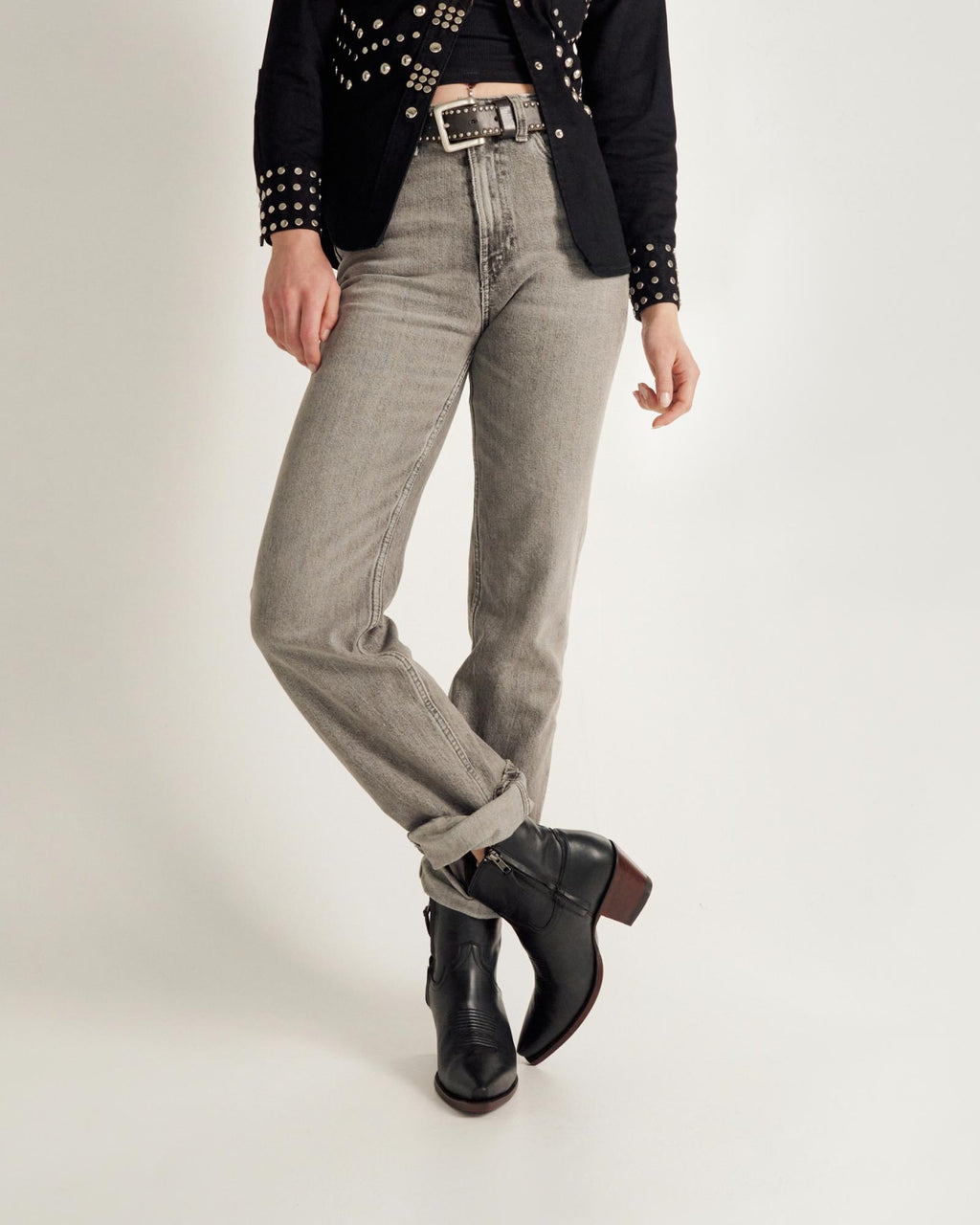 Person wearing light gray jeans, black ankle boots, a black belt with silver details, and a black studded jacket, standing against a plain white background.