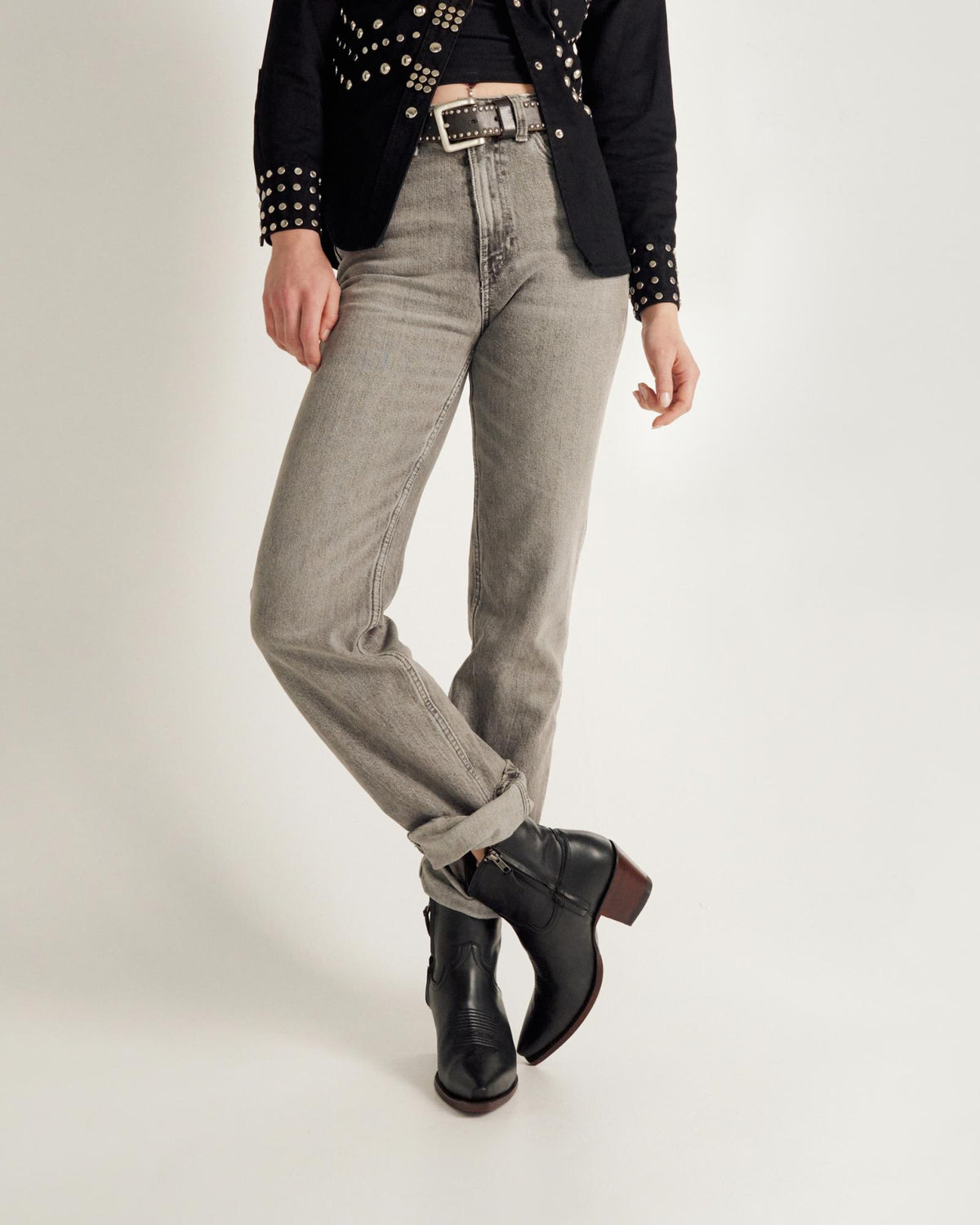 Person wearing light gray jeans, black ankle boots, a black belt with silver details, and a black studded jacket, standing against a plain white background.