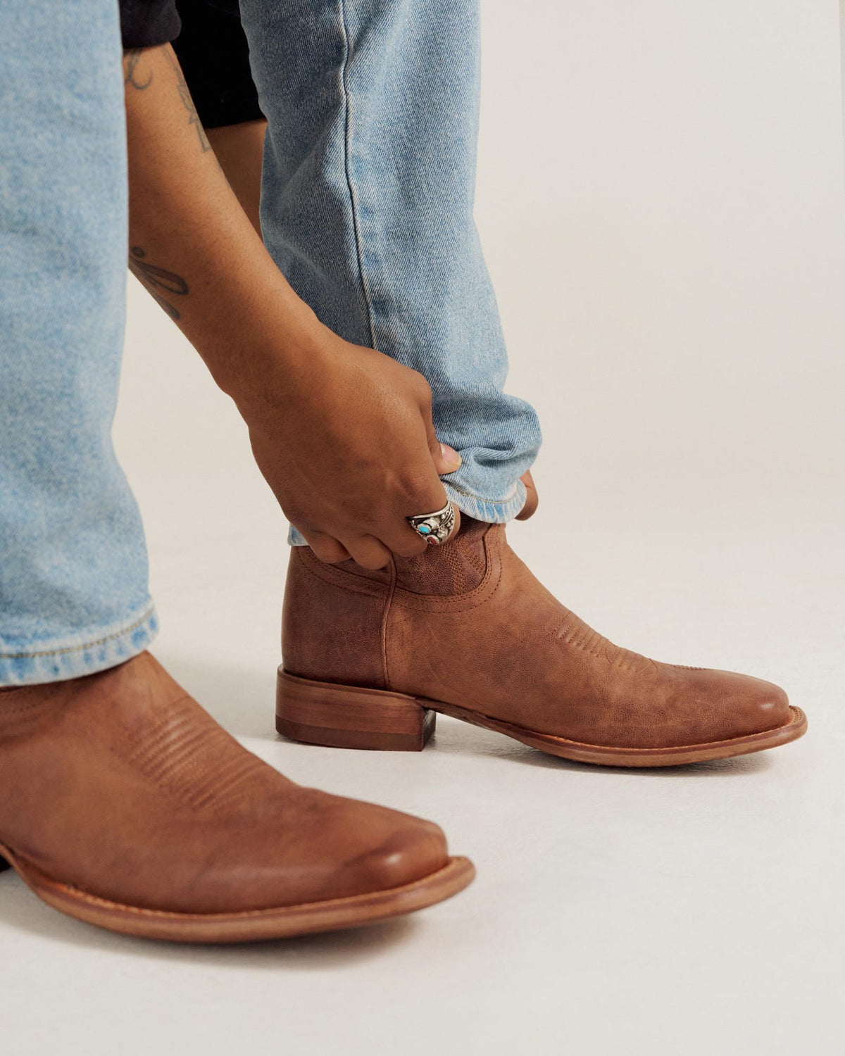 A person wearing light blue jeans adjusts the cuff of their pant leg above a brown leather ankle boot.