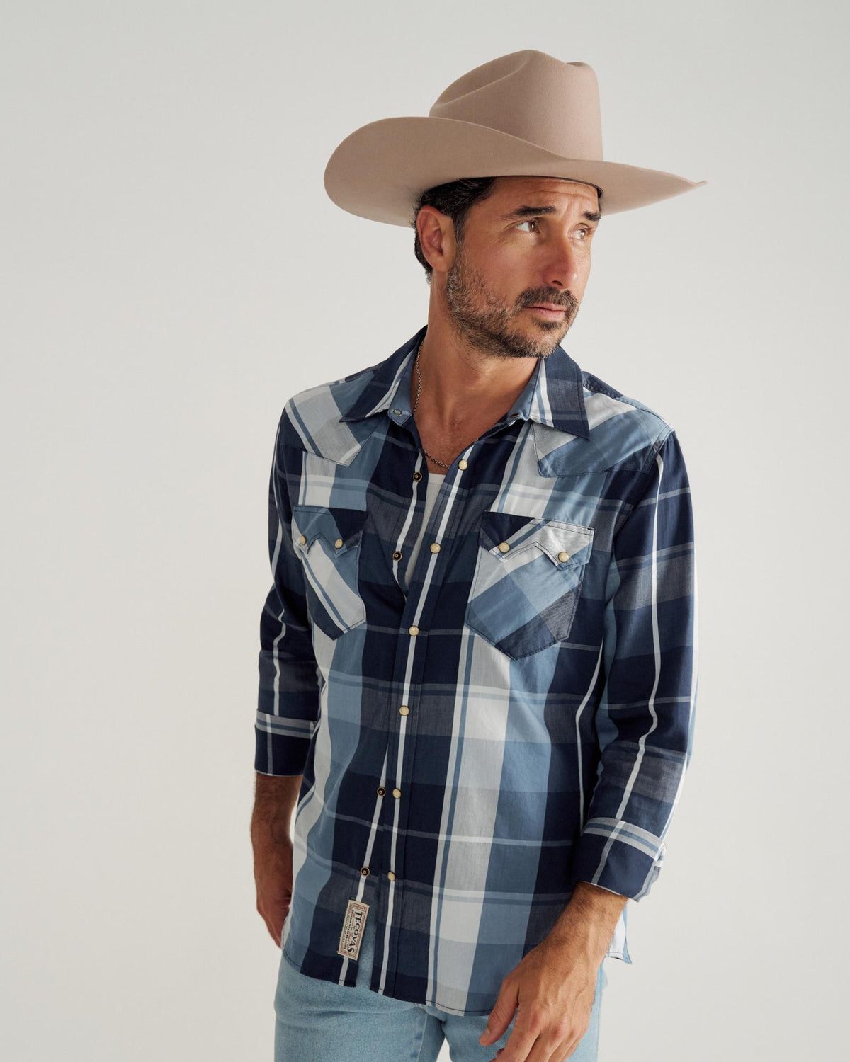 Man wearing a beige cowboy hat, blue plaid shirt, and light blue jeans stands against a plain white background, looking off to the side.