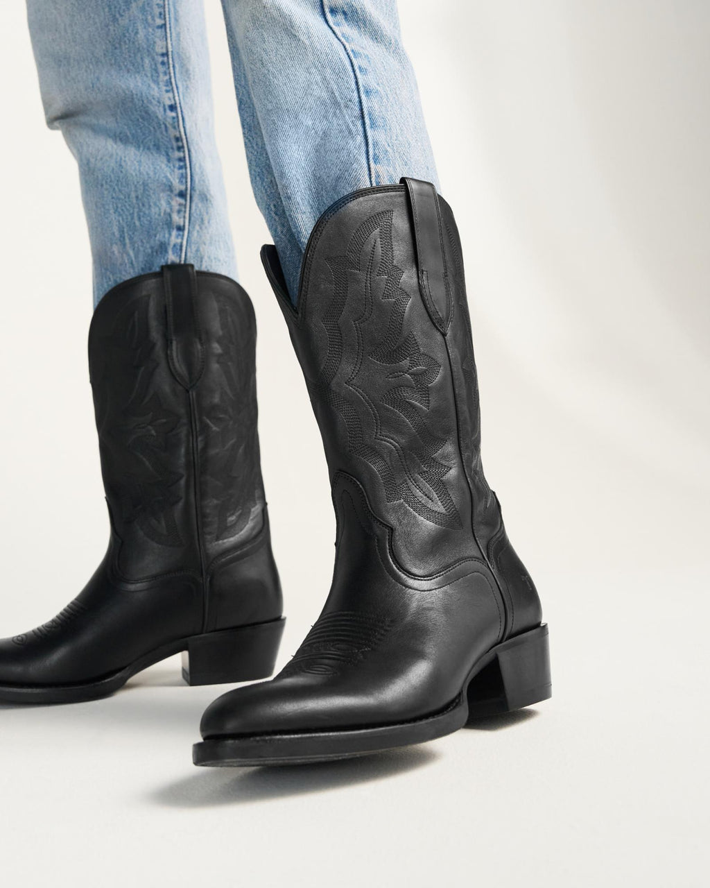 The Britt in Midnight Cowhide. A person wearing light blue jeans and black leather cowboy boots with decorative stitching in midnight cowhide stands on a white surface.