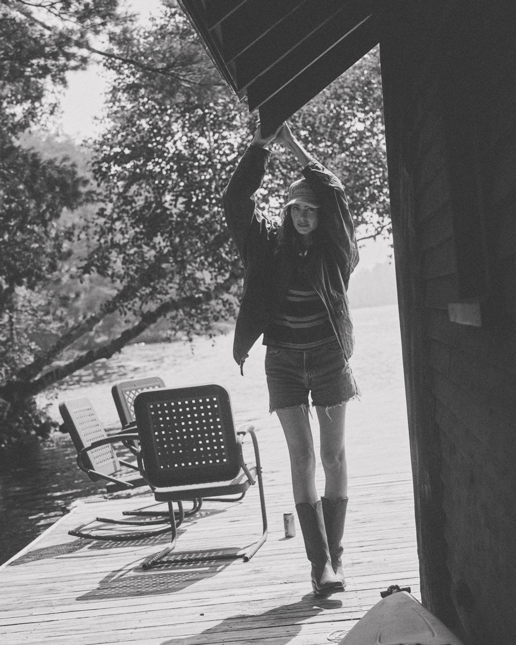 The Charlie in Scarlet Cowhide. A person in boots and a jacket stands on a lakeside dock, holding onto the roof edge. Scarlet Cowhide chairs and trees are visible in the background.