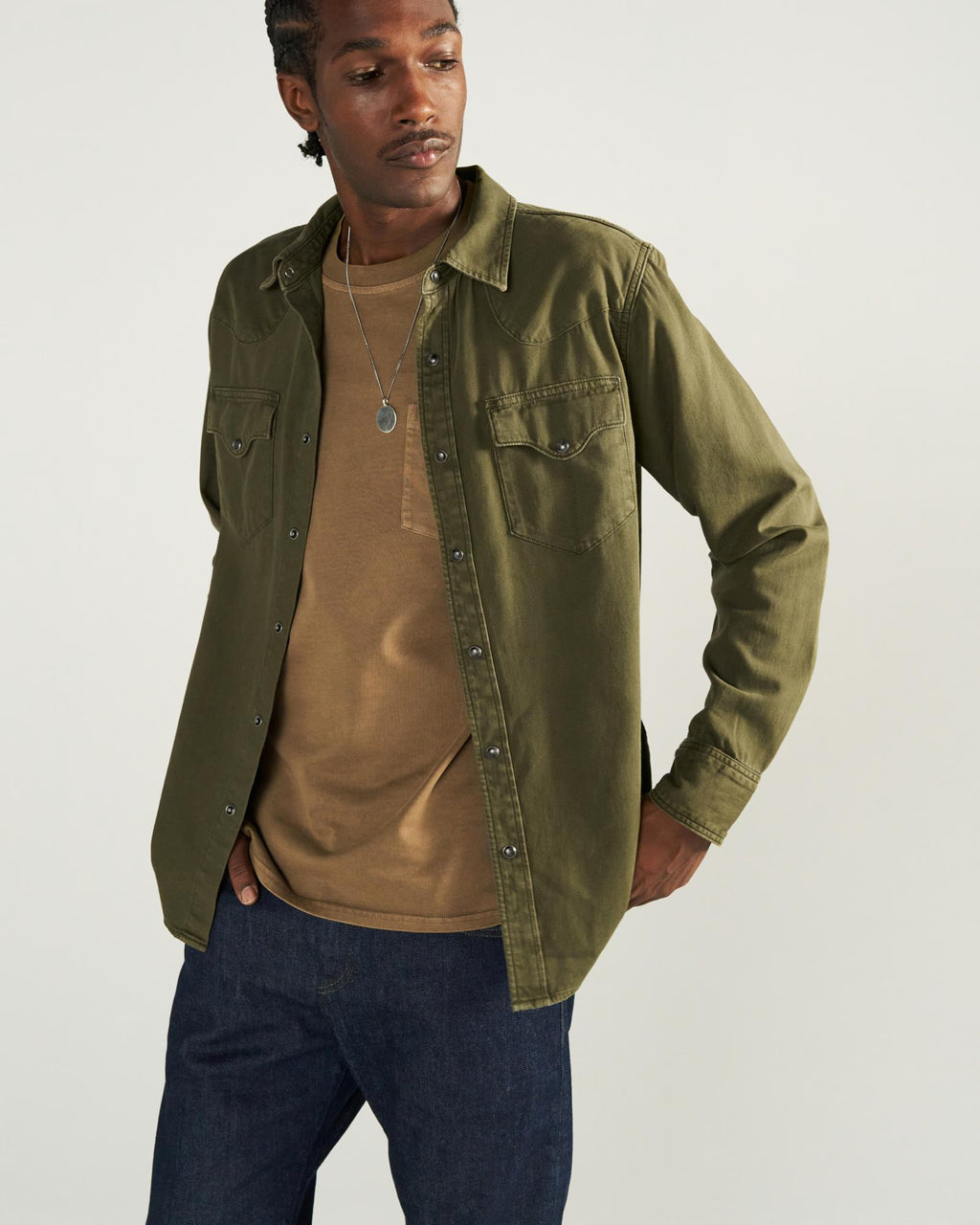 A man with a thin mustache and braided hair wears an olive green button-down shirt over a tan pocket t-shirt, dark blue jeans, and a silver pendant necklace against a plain light background.
