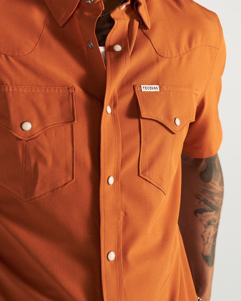 Person wearing an orange short-sleeve button-up shirt with two chest pockets and visible tattoos on their arm.