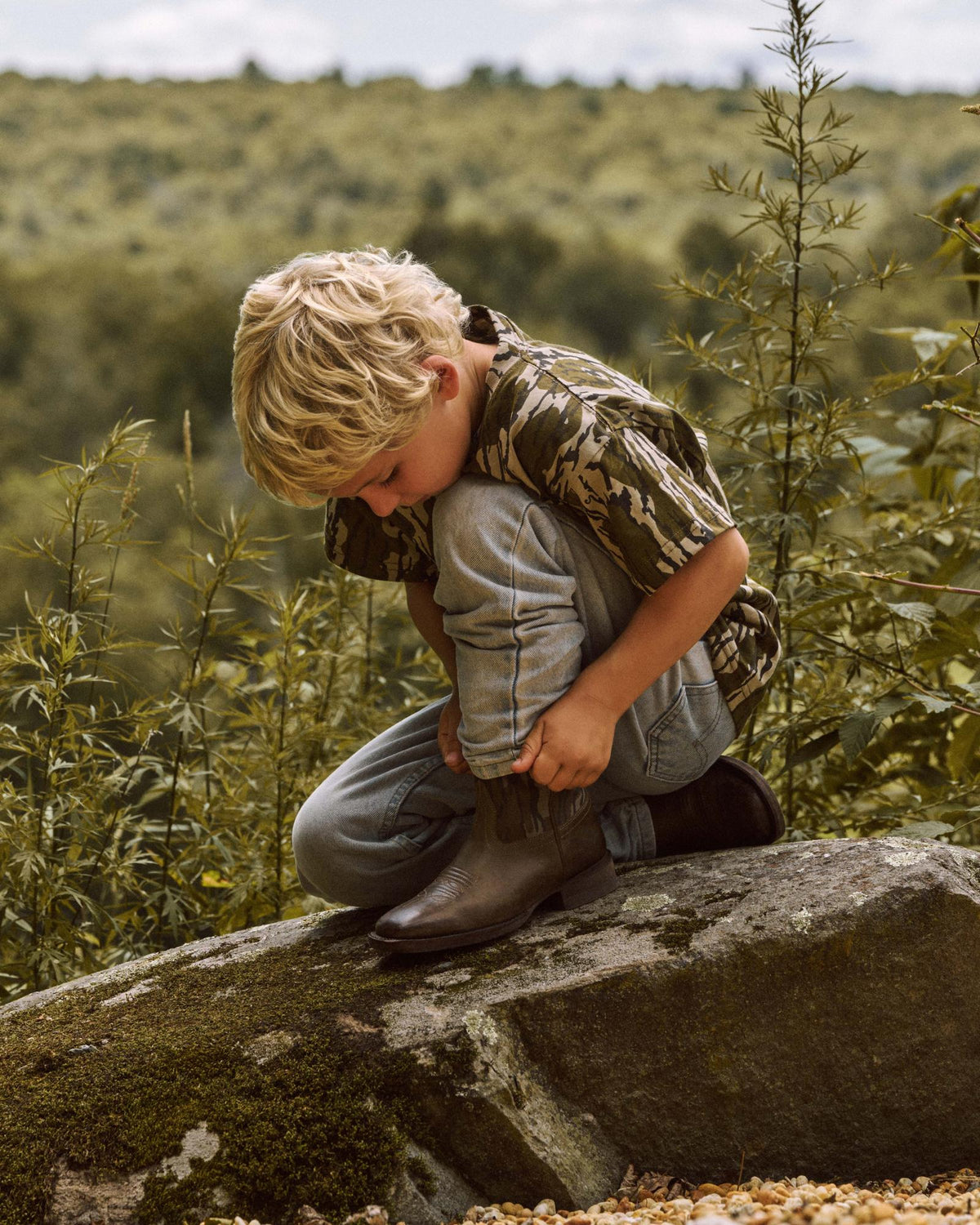 A young boy with blond hair crouches on a large rock outside, tying the laces of his brown boots.