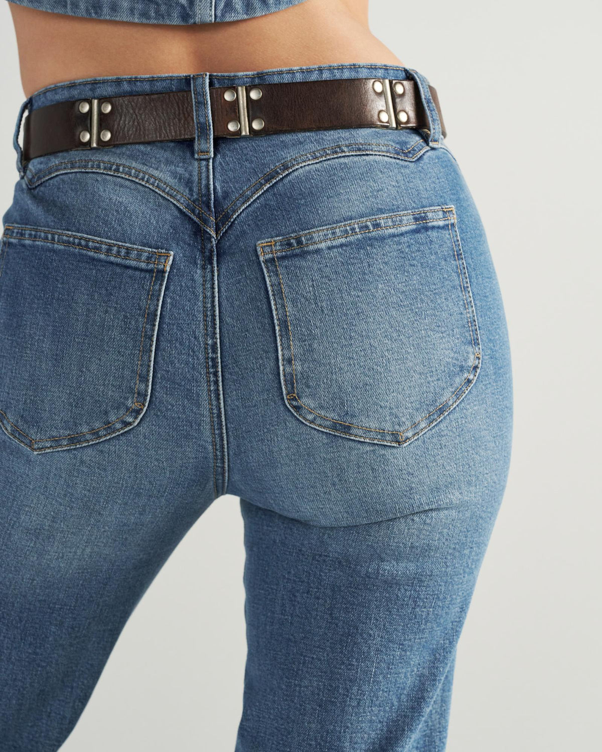 Person wearing blue jeans and a dark brown belt, shown from the waist down and viewed from the back against a plain background.