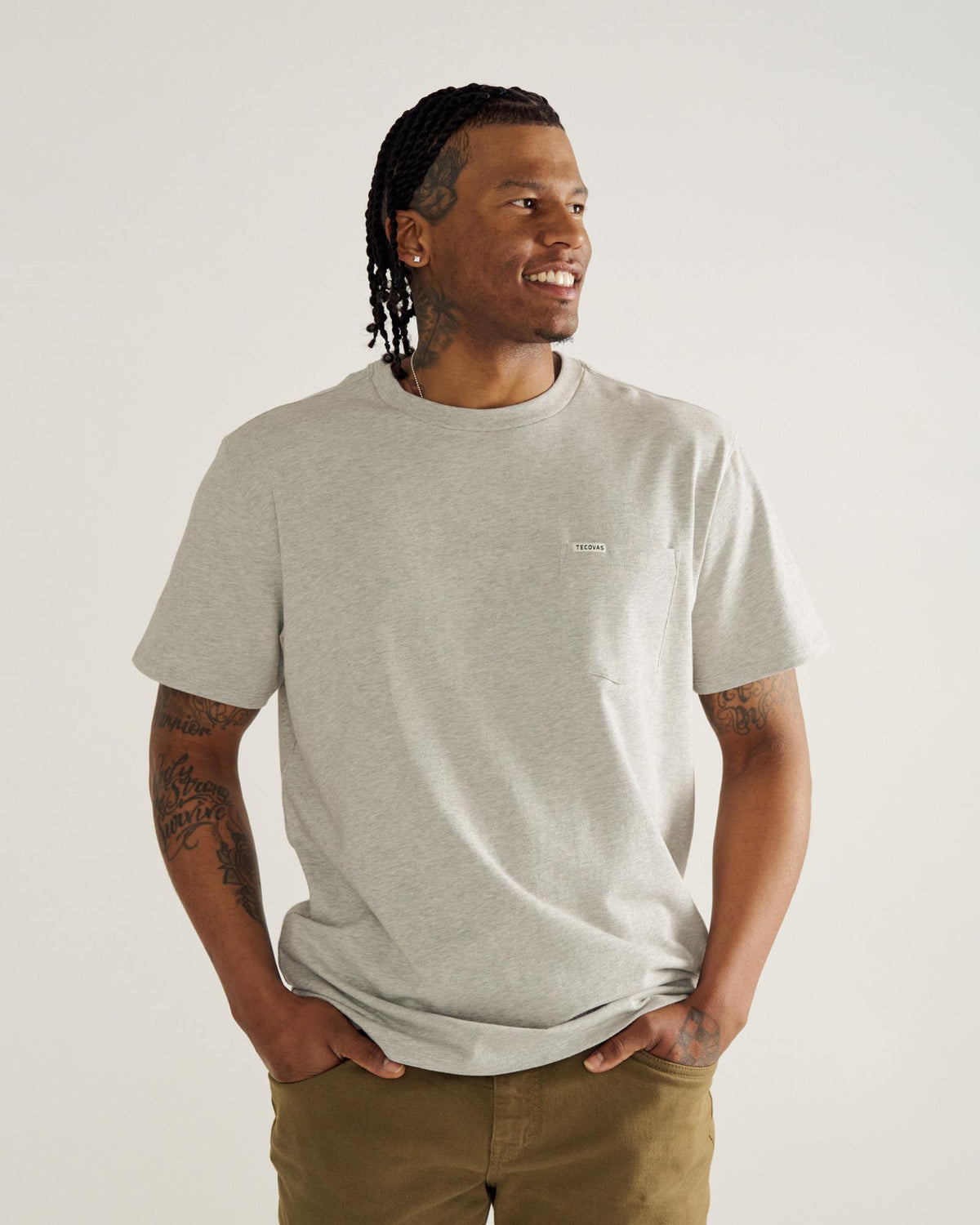 A man with braided hair and tattoos wears a light gray t-shirt and khaki pants, standing against a plain background with his hands in his pockets, looking to the side and smiling.