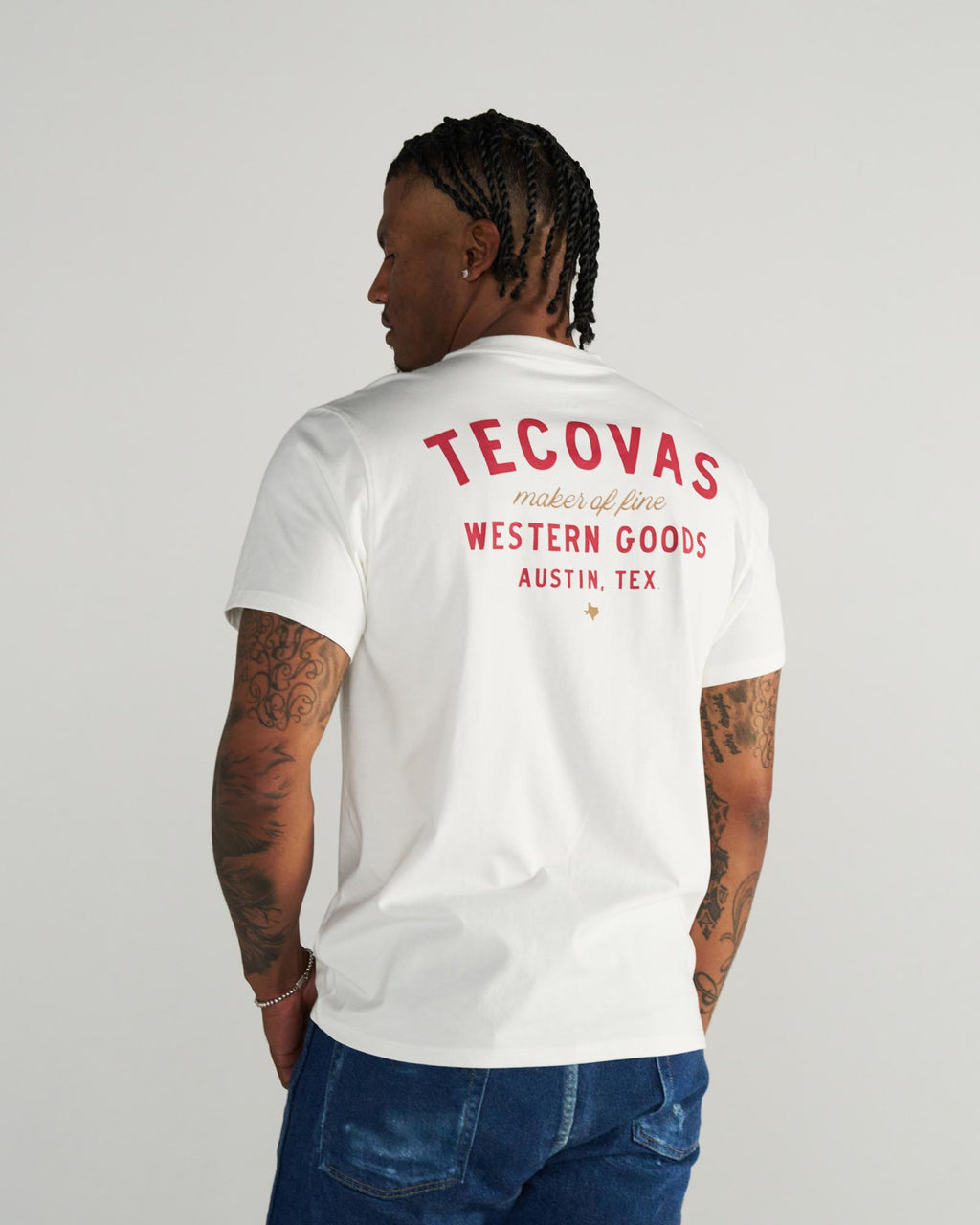 A person with braided hair wears a white T-shirt with red text reading “Tecovas maker of fine Western Goods Austin, Tex” on the back, standing against a plain background.