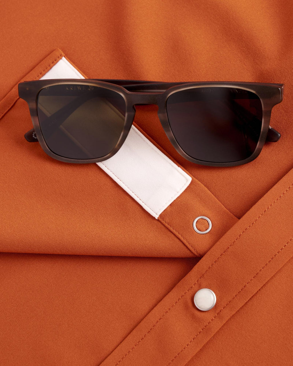 Black sunglasses rest on an orange shirt with a white pocket flap and silver snap buttons.