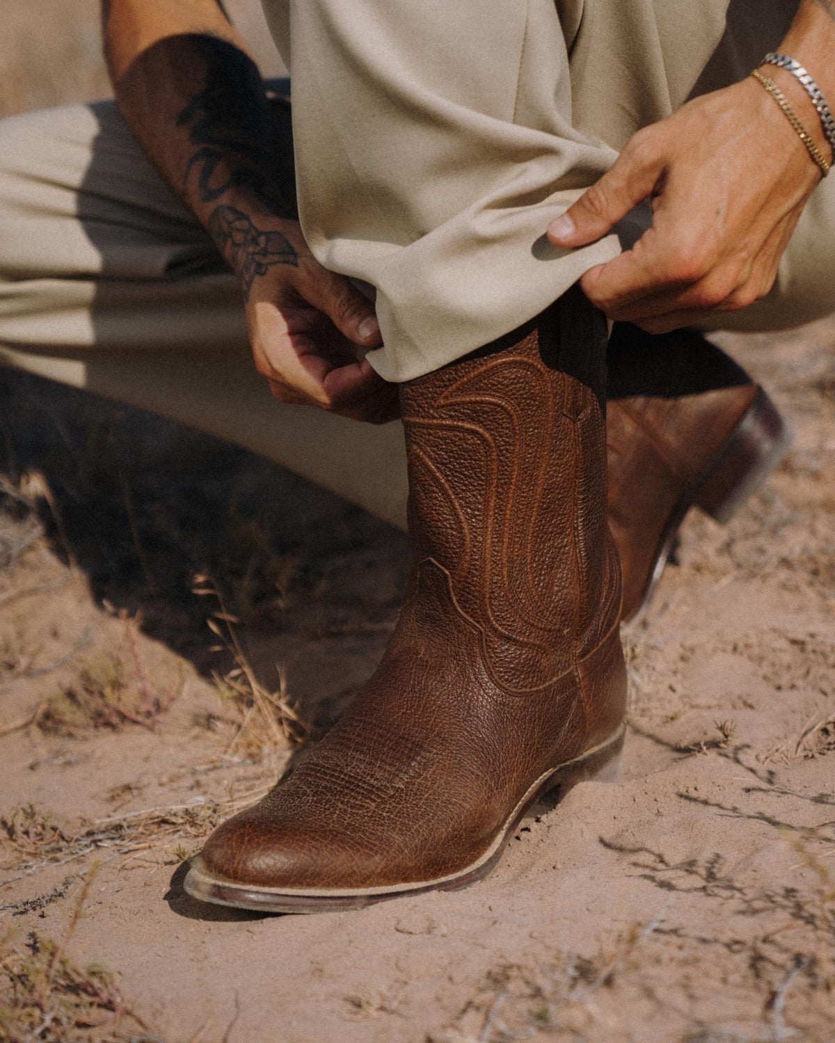 The Cartwright in Tobacco Regenerative Bison. A person wearing light pants adjusts their pant leg to reveal a brown leather cowboy boot while crouching on dry, grassy ground—a scene reminiscent of ranch life at The Cartwright Regenerative Bison.